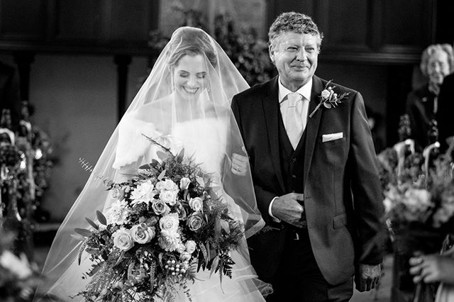 A bride walks down the aisle with her father, carrying a bouquet, in an elegant black and white wedding photograph.