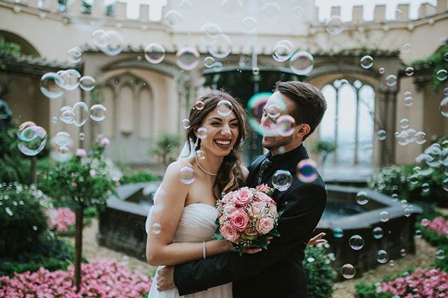 A smiling bride and groom share a loving embrace amidst a flurry of bubbles in a romantic courtyard setting.