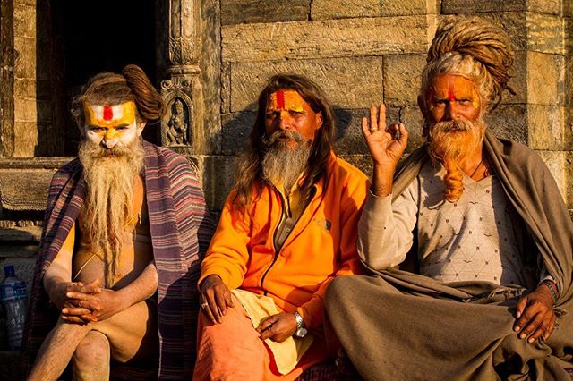 Three hindu monks with traditional face paint and long beards sit outside near a stone wall.