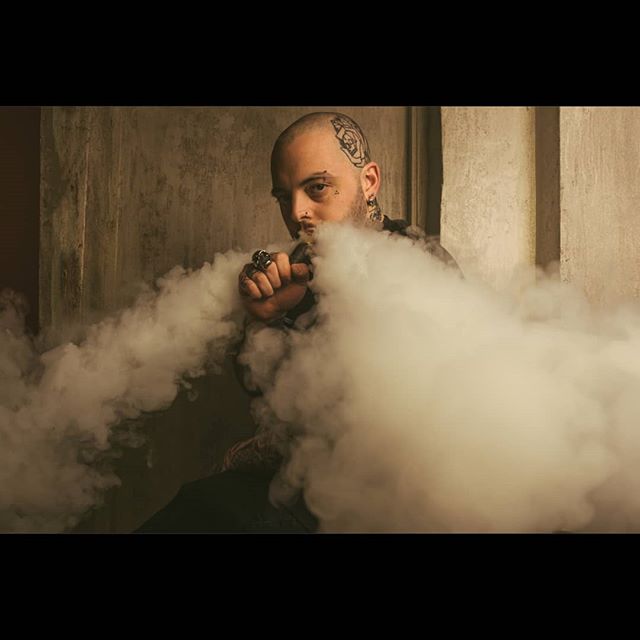 A tattooed man dramatically exhales a thick cloud of vapor in a moody indoor setting.
