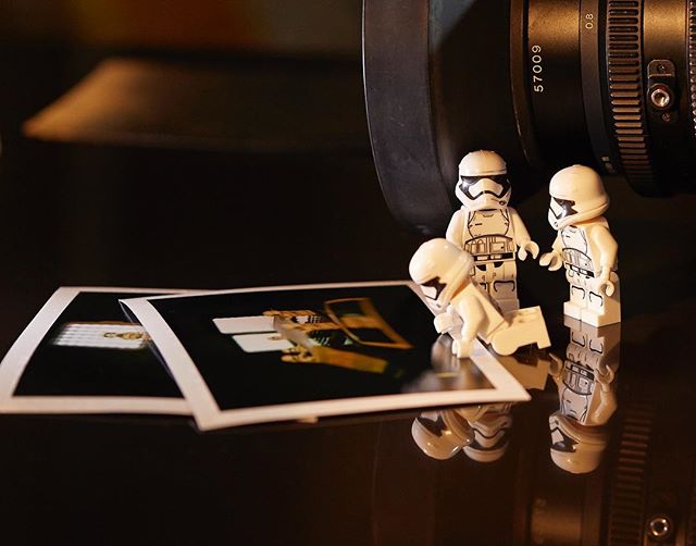 Three Lego stormtroopers examine printed photographs next to a camera lens on a reflective surface.