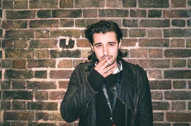 A man with dark hair stands in front of a brick wall, smoking a cigarette in a moody setting.