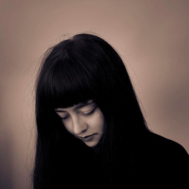 A contemplative woman with dark hair gazes downward in a close-up portrait with soft, muted tones and shallow depth of field.