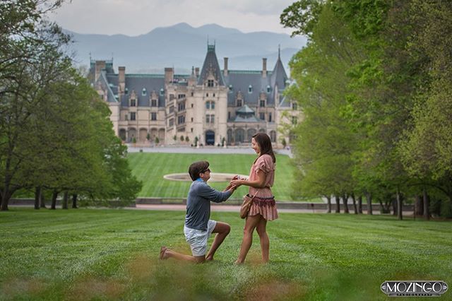 A man proposes to a woman in front of Biltmore Estate on a grassy lawn on a cloudy day.