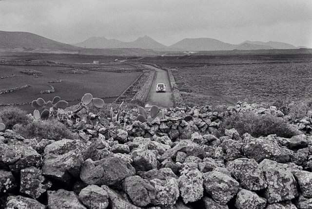 A car drives along a rural road lined with stone walls, set against a backdrop of mountains in a black and white landscape.