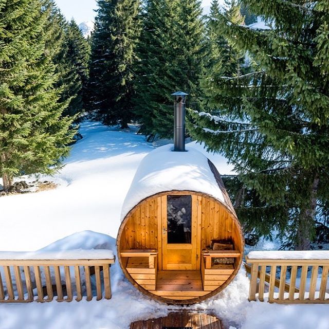 A cozy barrel sauna sits in a snowy winter landscape surrounded by evergreen trees, offering a tranquil escape.