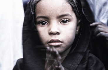 A young girl with expressive eyes looks directly at the camera with a serious expression in a close-up portrait.
