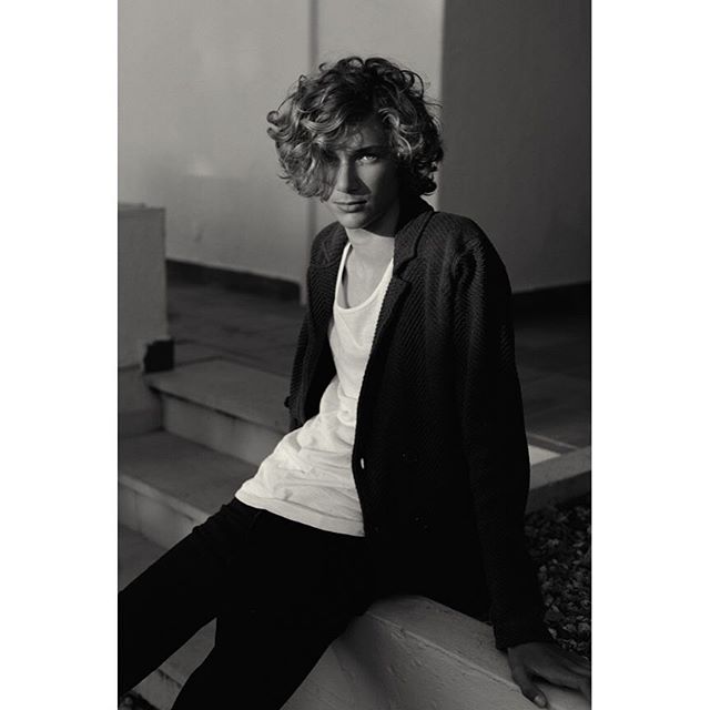 A young man with curly hair poses in a black and white shot, exuding confidence and a relaxed vibe.