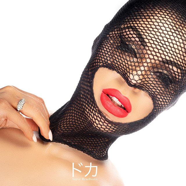 A woman in a black fishnet face mask with bold red lipstick poses for a striking studio shot.