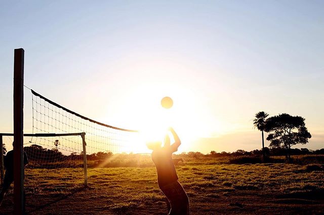 A volleyball player serves the ball in silhouette against a bright sunset sky on an outdoor court.