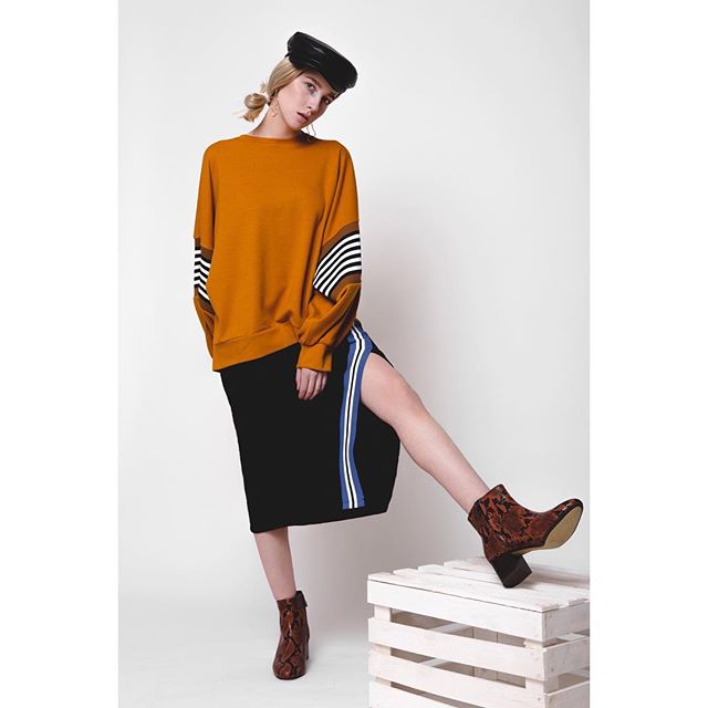 A fashionable woman poses in a mustard sweater, black skirt, hat and brown boots. 