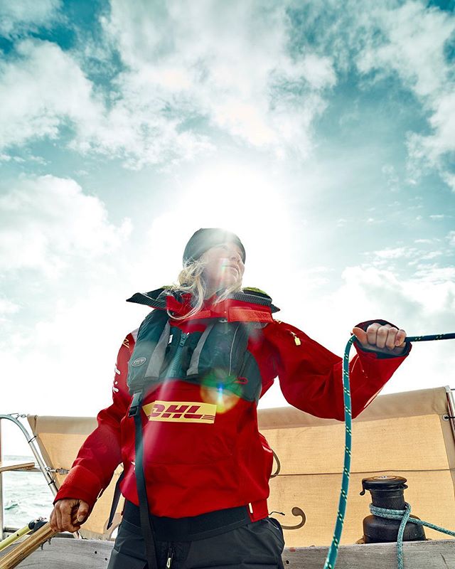A determined woman sailing on a sailboat, wearing a DHL branded jacket, enjoying a sunny day on the sea.