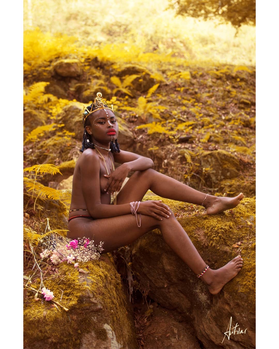 A nude woman with tribal jewelry sits in a golden forest, creating a dreamy and sensual atmosphere.