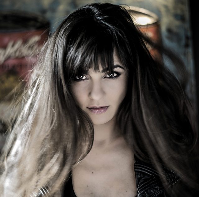 Intense brunette woman with bangs stares directly at the camera in a moody, close-up portrait.