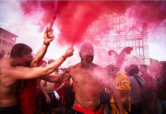 A lively crowd of shirtless men celebrates in the city with red smoke bombs during a cultural event.