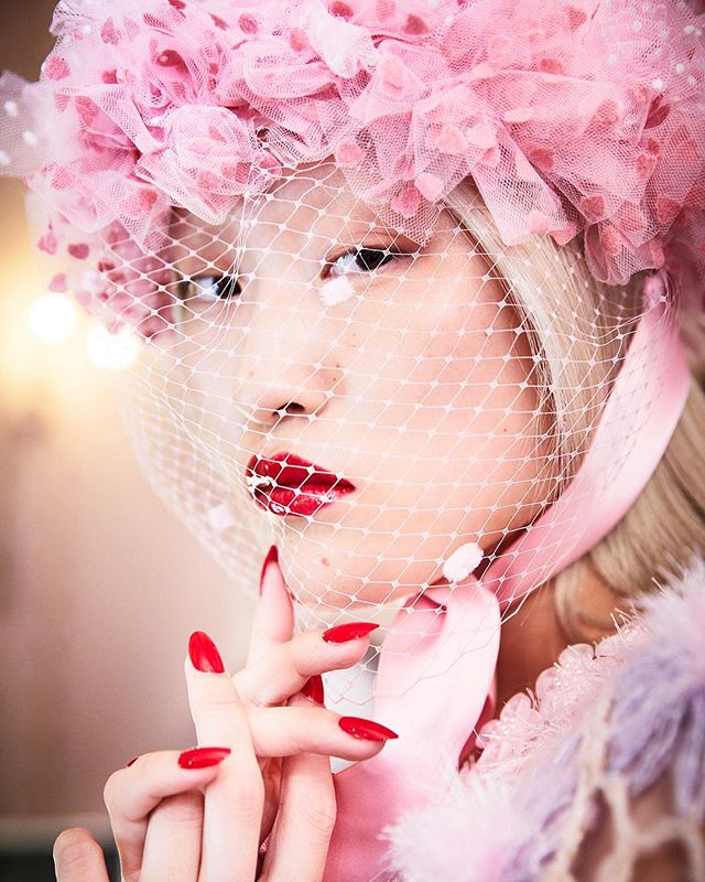 A woman with red lipstick and nail polish wears a pink headpiece with a veil in a glamour shot.