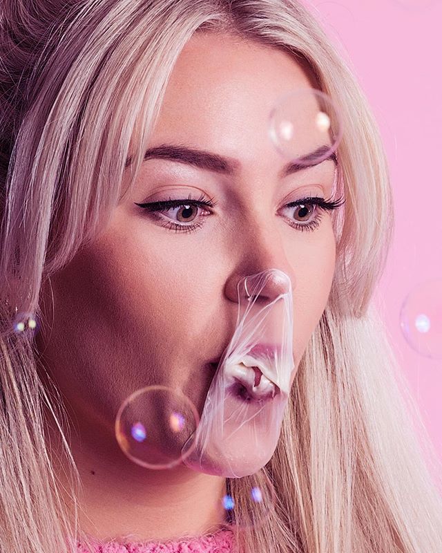 A blond woman playfully blows chewing gum bubbles against a vibrant pink backdrop, perfect for beauty campaigns.