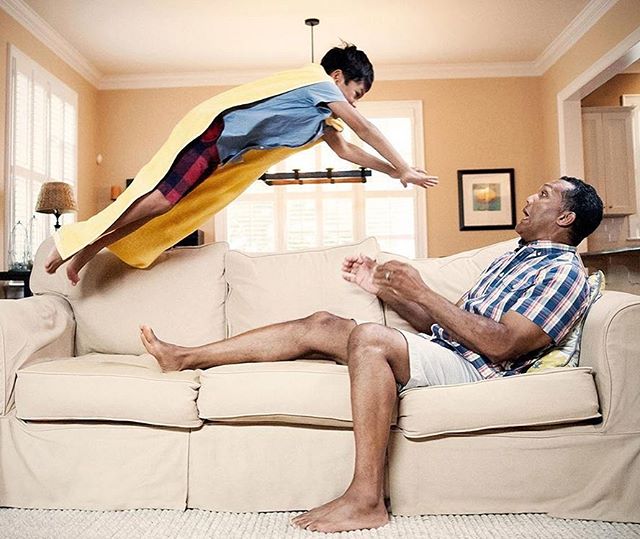 A boy wearing a yellow blanket as a cape jumps toward his father who is relaxing on a beige couch in their living room.