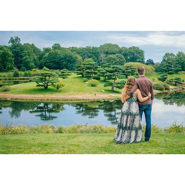 A couple stands embraced, gazing at a serene lake surrounded by lush greenery and manicured trees.