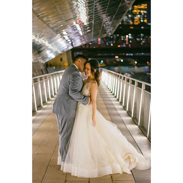 A bride and groom embrace on a bridge at night, with a cityscape in the background.