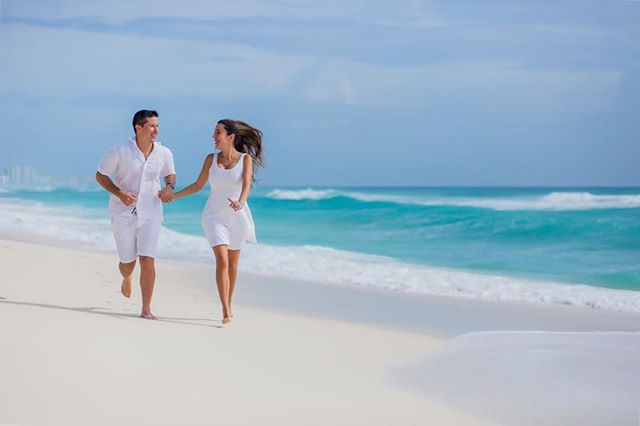A smiling couple in white runs hand-in-hand along a sunny beach next to the ocean.