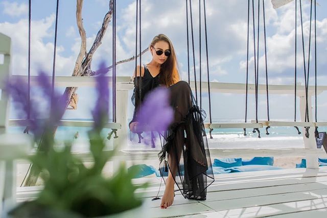 A woman in a black dress and sunglasses sits on a swing at the beach.