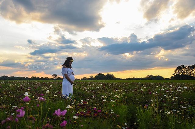 A pregnant woman in a white dress stands in a field of flowers at sunset.