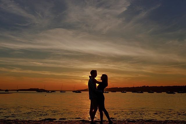 A silhouetted couple embrace on a beach at sunset, bathed in warm, romantic light against a backdrop of boats and a serene ocean.