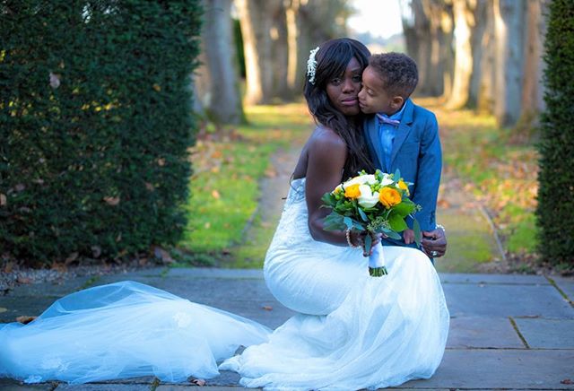A bride in a white dress kneels on a stone path, holding a bouquet, while a young boy kisses her cheek in a loving moment.