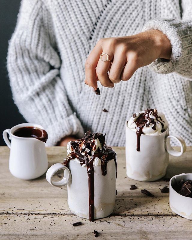 A hand sprinkles chocolate over mugs of hot chocolate topped with cream and chocolate sauce on a rustic wooden table.