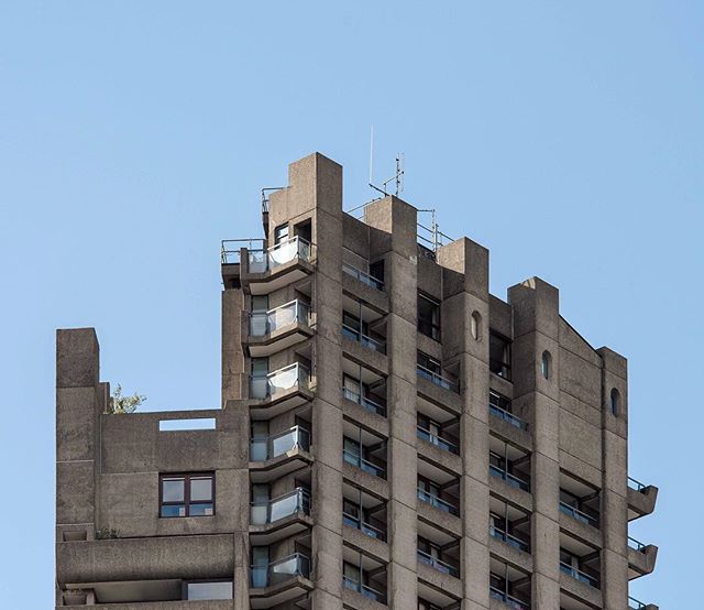 A low angle shot shows the top of a concrete, brutalist high-rise building against a clear blue sky.
