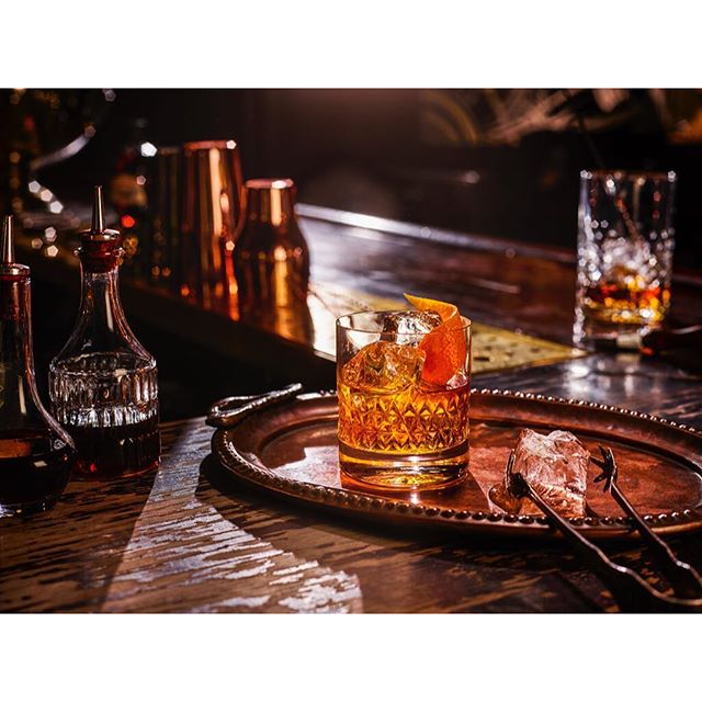 A glass of old fashioned cocktail with ice and orange peel on a copper tray in a bar.