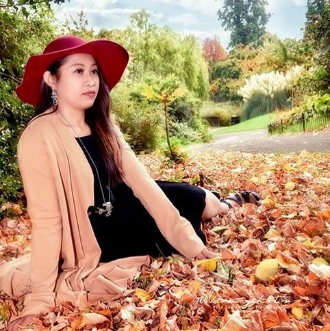 A serene woman in a hat and cardigan poses among autumn leaves in a park. 