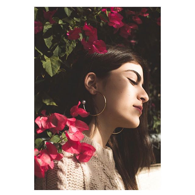 A woman with hoop earrings poses among bright pink bougainvillea flowers in soft, natural light.