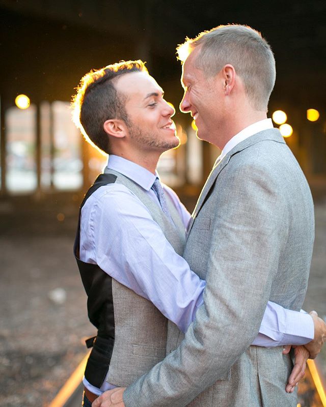 A smiling gay couple embraces warmly on railroad tracks at golden hour for a portrait of love.