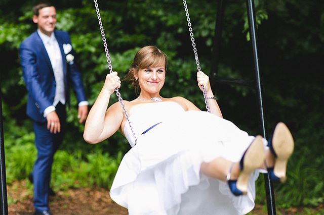 A bride enjoys a swing on her wedding day with the groom in the background.