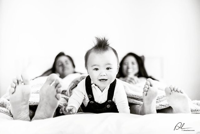 A baby crawls toward the camera with their parents lying behind them in bed in a black and white photo.