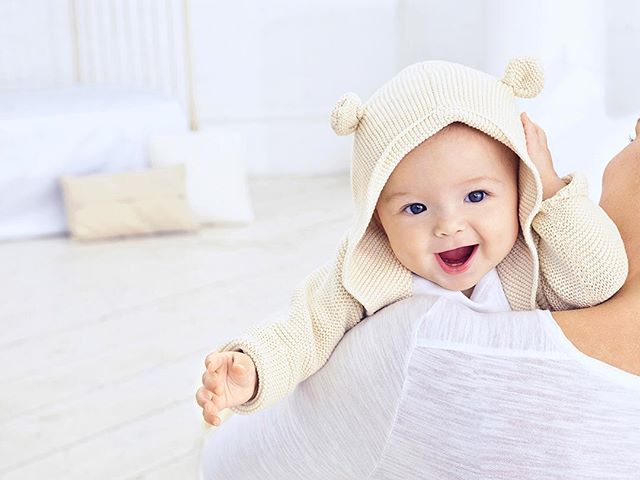 Adorable baby with blue eyes smiles while being held. The baby is wearing a knitted hoodie.