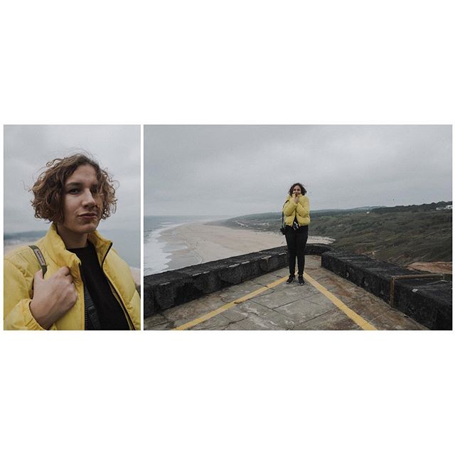 A woman in a yellow jacket stands on a cliff overlooking the beach and ocean on an overcast day.