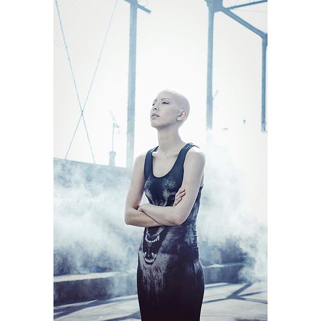 A bald woman in a wolf print dress stands outdoors with arms crossed in an urban setting.