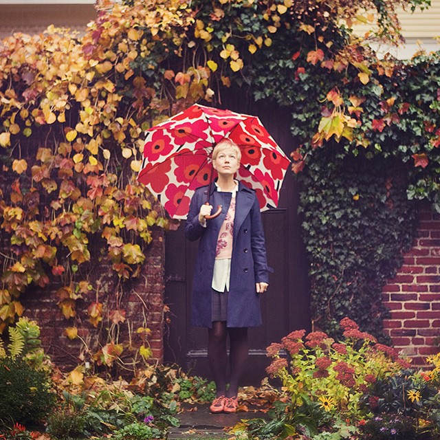 A woman stands with an umbrella in a garden with autumn foliage, creating a serene and seasonal scene.