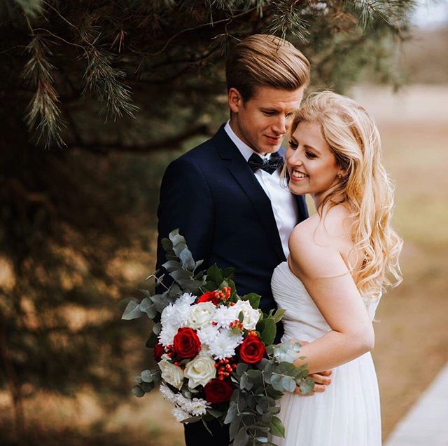 A happy bride and groom embrace, holding a bouquet of red and white roses on their wedding day.