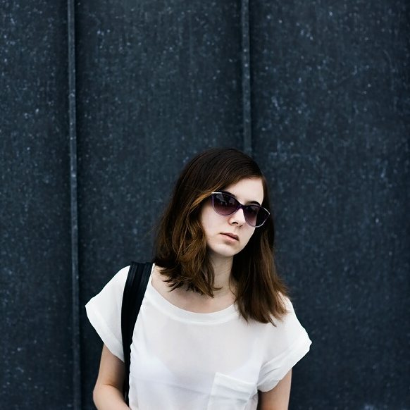 A stylish brunette woman with sunglasses poses against an urban backdrop, capturing a moment of calm contemplation.