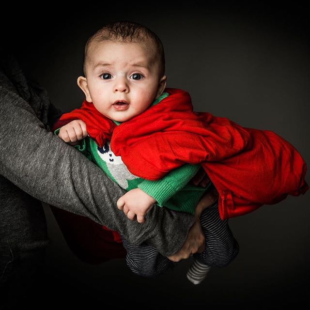 Adorable baby in a red cape and green sweater held in someone's arms. 
