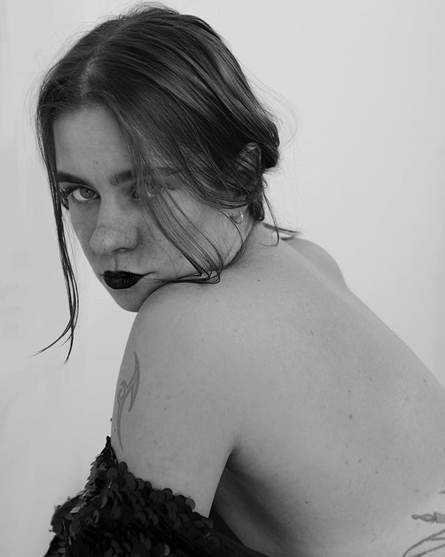 A woman with dark lipstick poses in a black and white portrait with a tattoo on her shoulder.