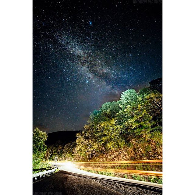 A winding road at night with car light trails under a starry Milky Way sky with green trees.