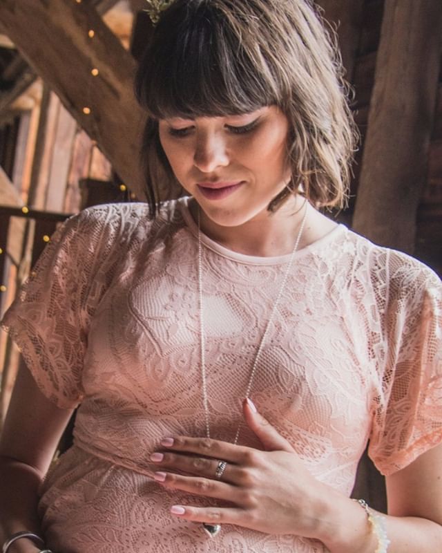 A pregnant woman in a pink lace dress gently places her hand on her belly.