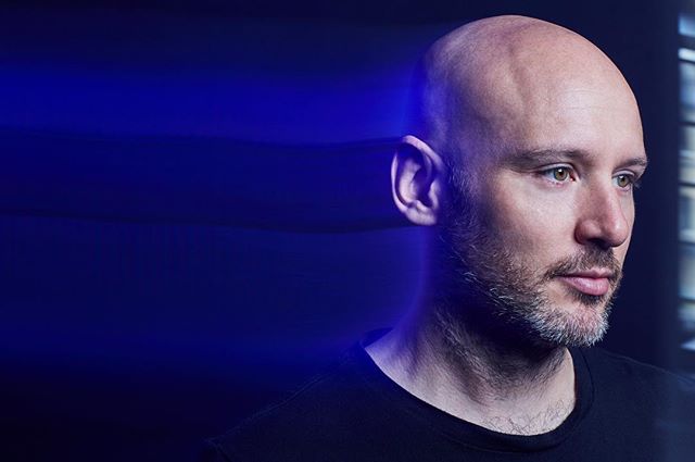 A bald man gazes intently in a dramatic, studio portrait with blue lighting and a modern style.