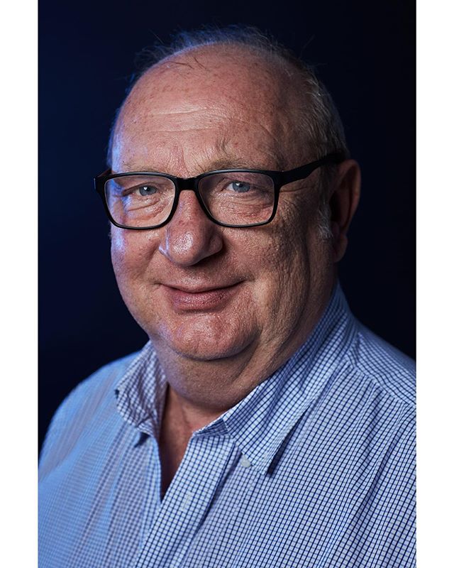 A confident man with glasses smiles in a professional studio portrait against a dark background.
