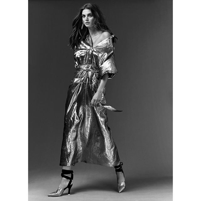 A stylish woman poses in a silver dress and heels in a black and white studio shot.